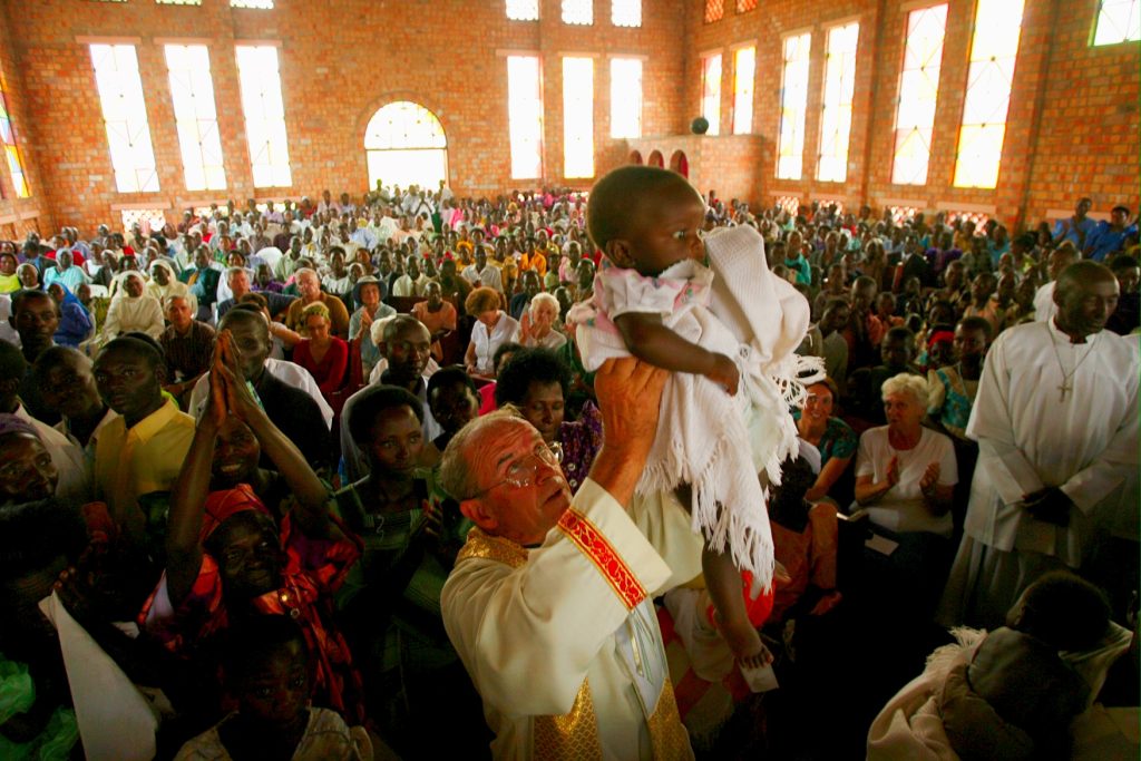 priest lifts baby up high with crowd in background